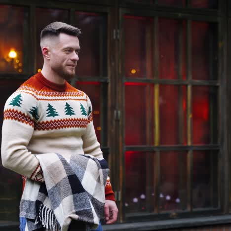 smiling young guy on cottage background. handsome man in warm sweater walking to someone in terrace. guy with a warm blanket in hands outdoors.