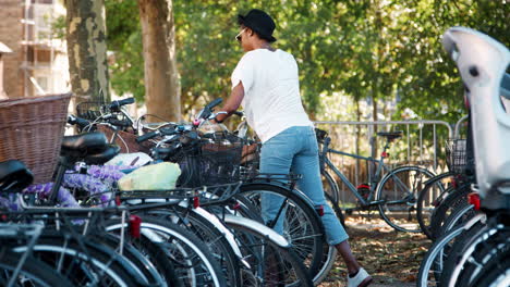 Young-black-woman-wearing-white-t-shirt,-blue-jeans-and-sunglasses-parking-her-bicycle,-putting-on-crossbody-handbag-and-walking-away-smiling,-handheld