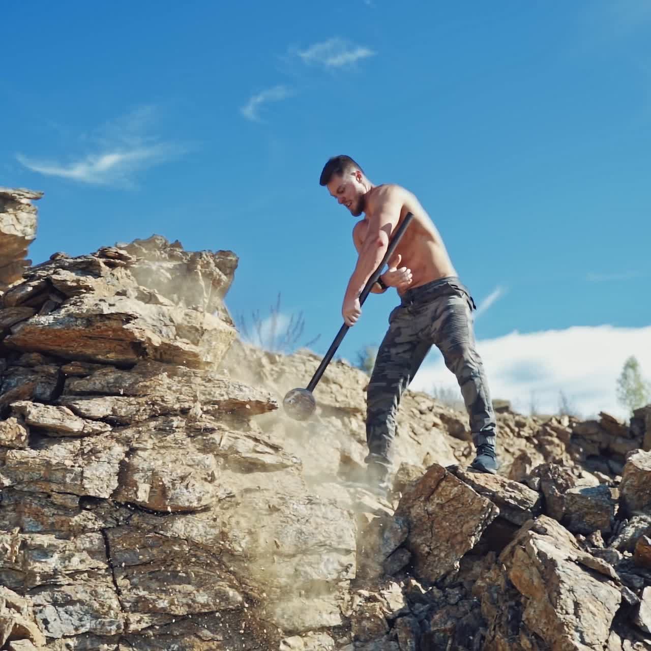 Muscular athlete with hammer on the rocks. Shirtless sportsman breaking stones with hard metal hammer outdoors. Slow motion.