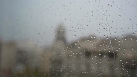 A-slow-motion-narrow-view-of-raindrops-seen-through-a-window-during-heavy-rainfall,-with-an-urban-city-landscape-in-the-background