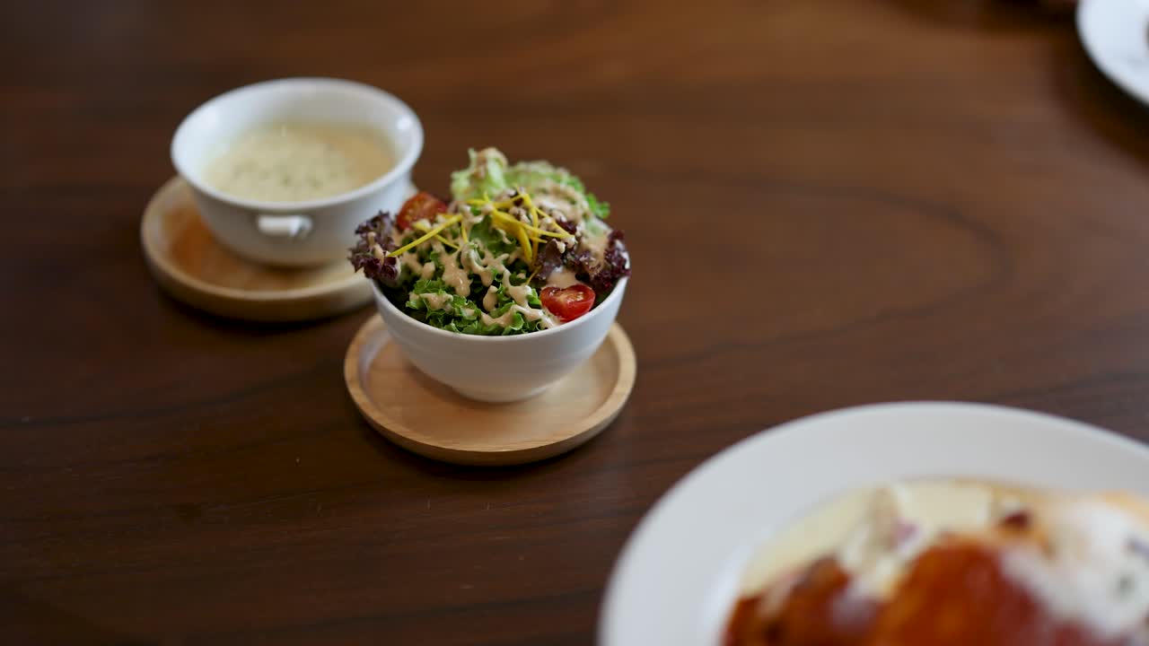 Hand places salad beside creamy omelette rice, soup on wooden table, natural light, static shot