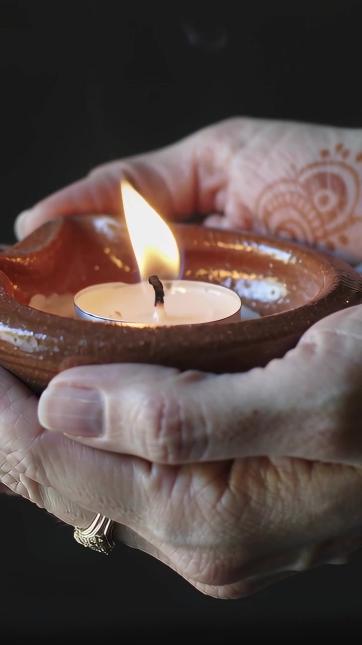 Vertical video: Cupping senior hands guarding clay diya in dark room, wick burning, henna
