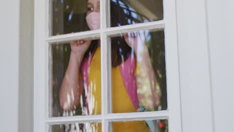 chica caucásica con máscara facial y mochila mirando por la ventana en casa