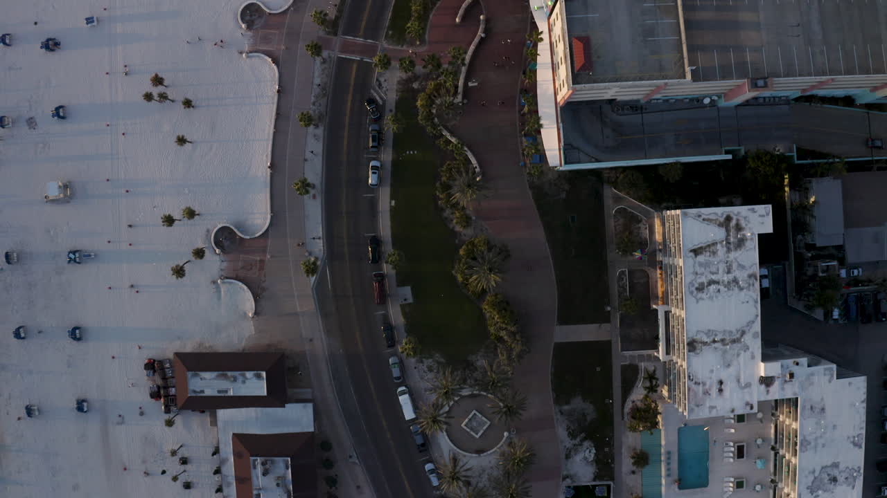 A direct overhead perspective reveals a beachfront boulevard lined with cars and palm trees, bordered by white sand on one side and hotel pools on the other