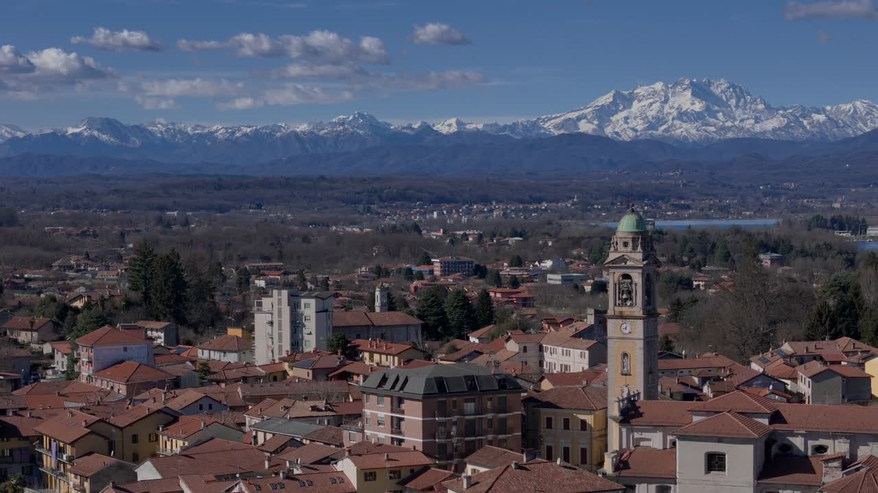 Aerial drone view of town with red-tiled roofs and clock tower, set against backdrop of snow-capped Alps and Lake Como Italy under clear blue sky on a bright daylight
