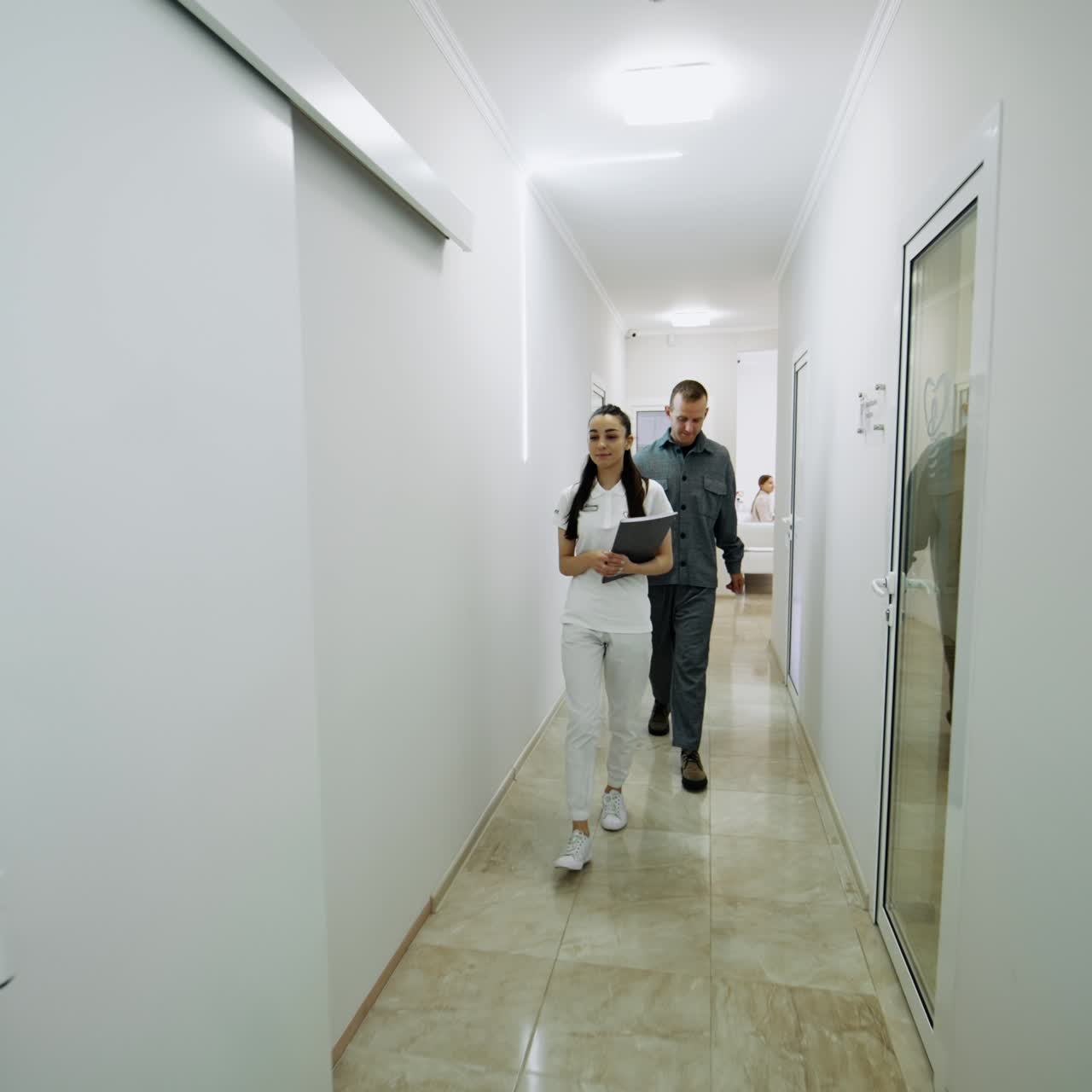 Positive attractive nurse taking patient to the doctor's cabinet. People walk a long white hall of a modern hospital
