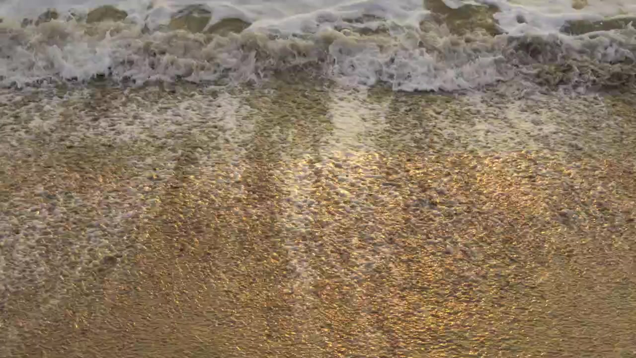 Foamy ocean water receding over wet sand with golden sunset reflections