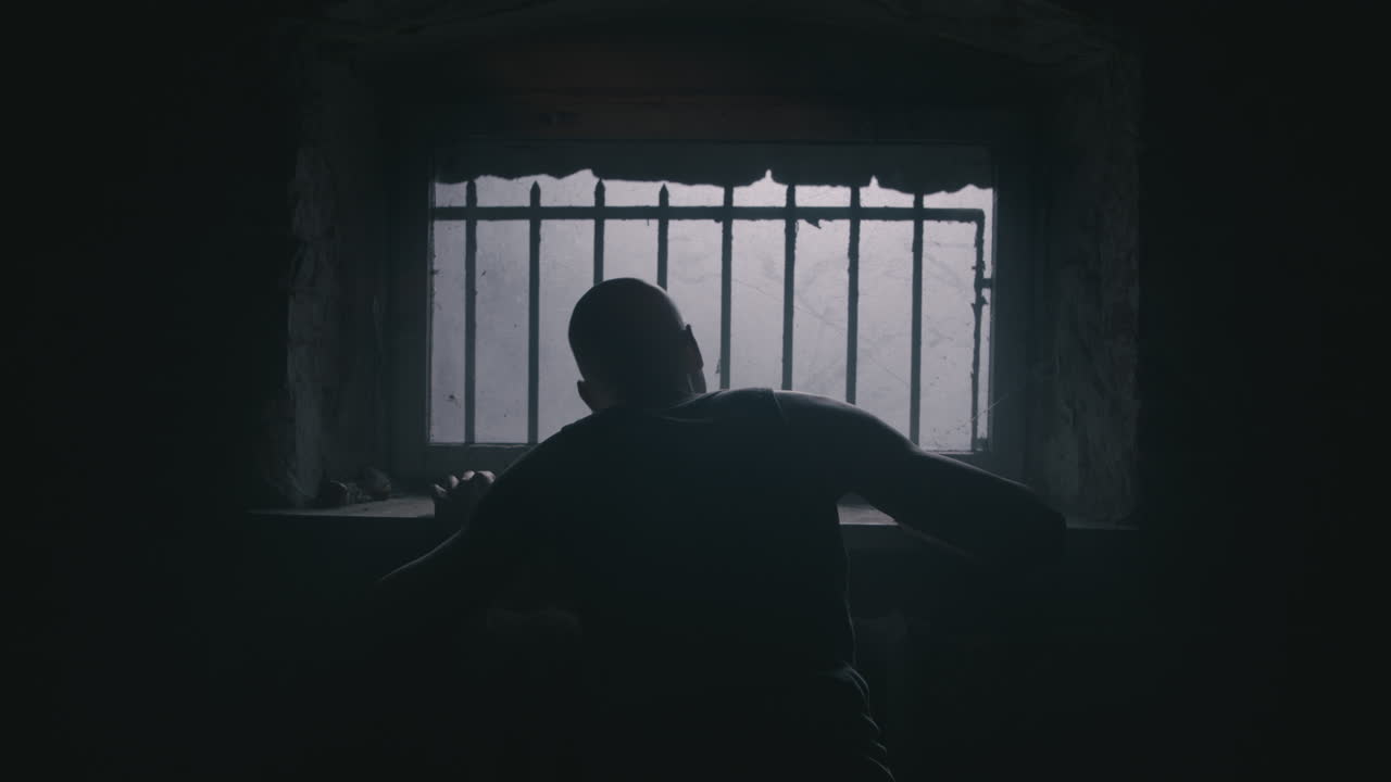 Pale bald skinny man looks through the window in the creepy old bathroom at night