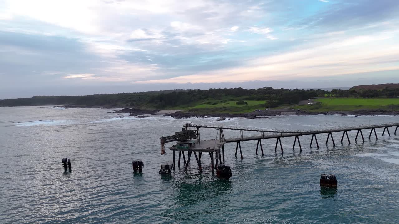 Vista aérea de órbita ampla de um cais industrial de carregamento de cascalho sob céus nublados, oceano calmo ao fundo