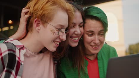 close-up shot of three happy girls with short long hair in bright clothes looking at something on a laptop screen during their walk in the summer