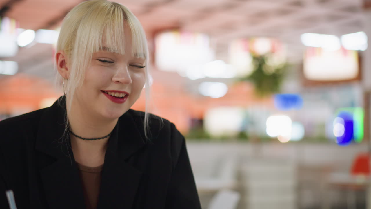 Young blonde lady in black jacket interacts with friend during conversation at bright indoor space, showing expression of focus and casual connection