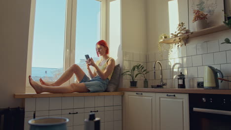 young woman sitting on sunlit windowsill in kitchen, texting on phone