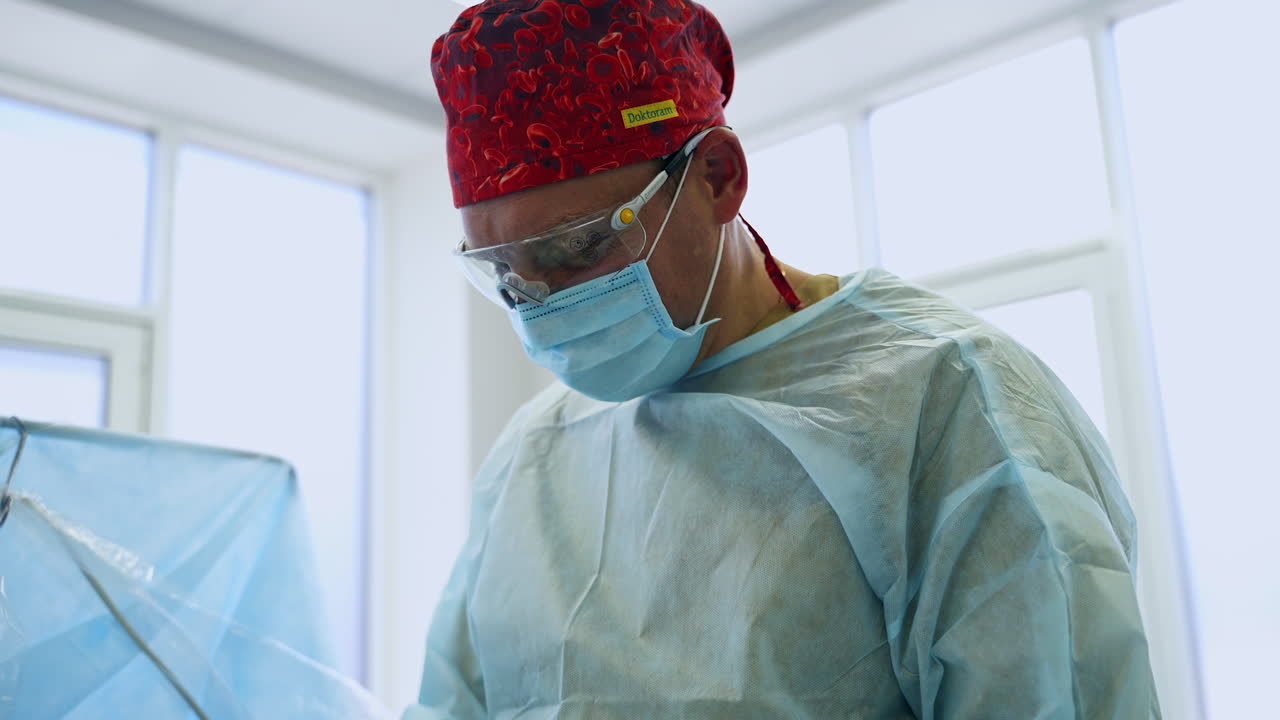 Adult male surgeon wearing uniform, red cap, mask and glasses. Medical professional being busy with operation. Low angle view.