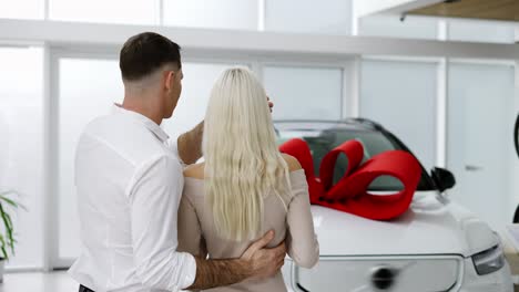 A-man-make-surprise-to-a-blonde-woman,-closed-woman's-eyes-from-back---new-car-with-red-bow,-rear-view