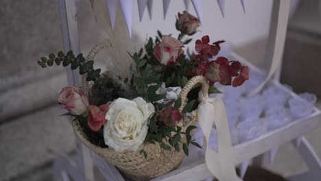 Cesta-Con-Rosas-Y-Vegetación-Atada-Con-Cinta-Sobre-Una-Silla-Decorativa-Blanca.