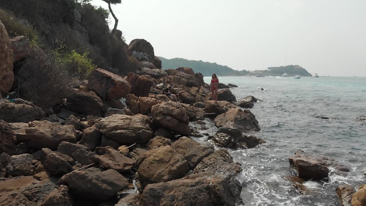 검은 머리카락의 여인이 파란 바다에 대항하는 거대한 돌 위에 서 있습니다.