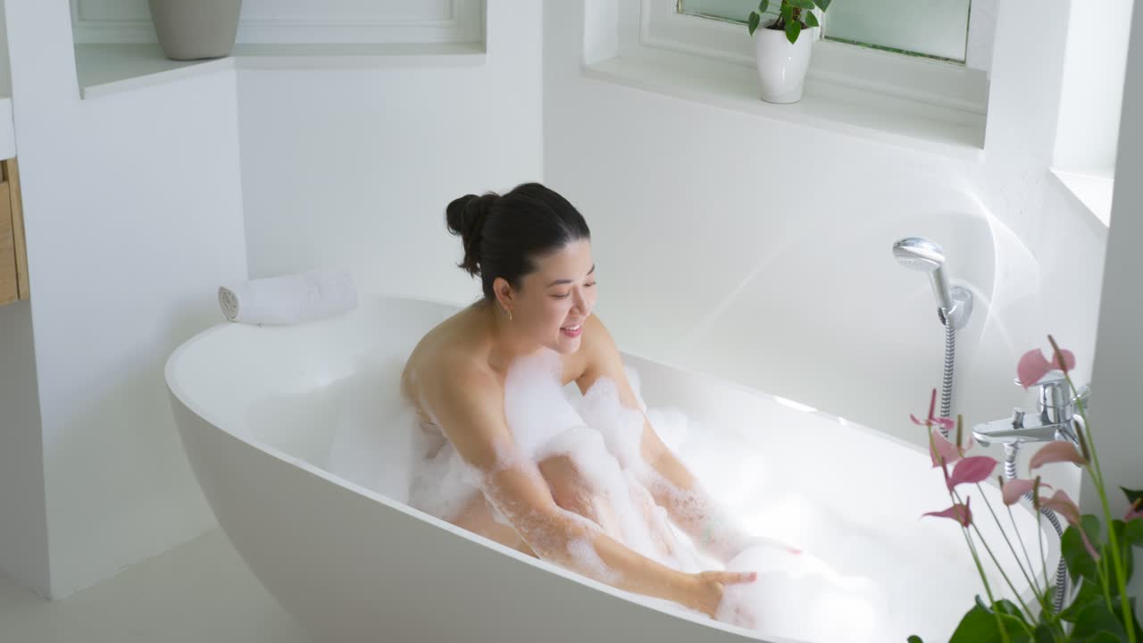 African American woman reaching forward, scooping foam in tub, blowing bubbles while relaxing