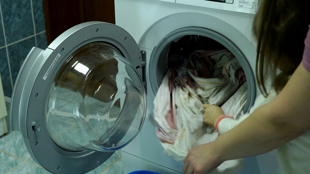 한 여성이 딸에게 세탁기에서 깨한 옷을 어떻게 가져올지 보여줍니다. 소녀는 집안에서 어머니를 돕는 법을 배니다. 비디오 촬영 클로즈업.