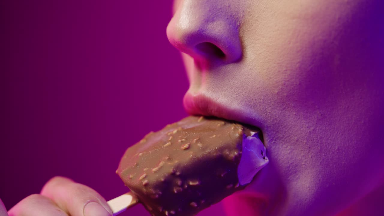 toma cercana de una mujer joven con hermosos labios rojos mientras come un helado de chocolate vegano en un palo y disfruta del sabor frente al fondo púrpura en cámara lenta