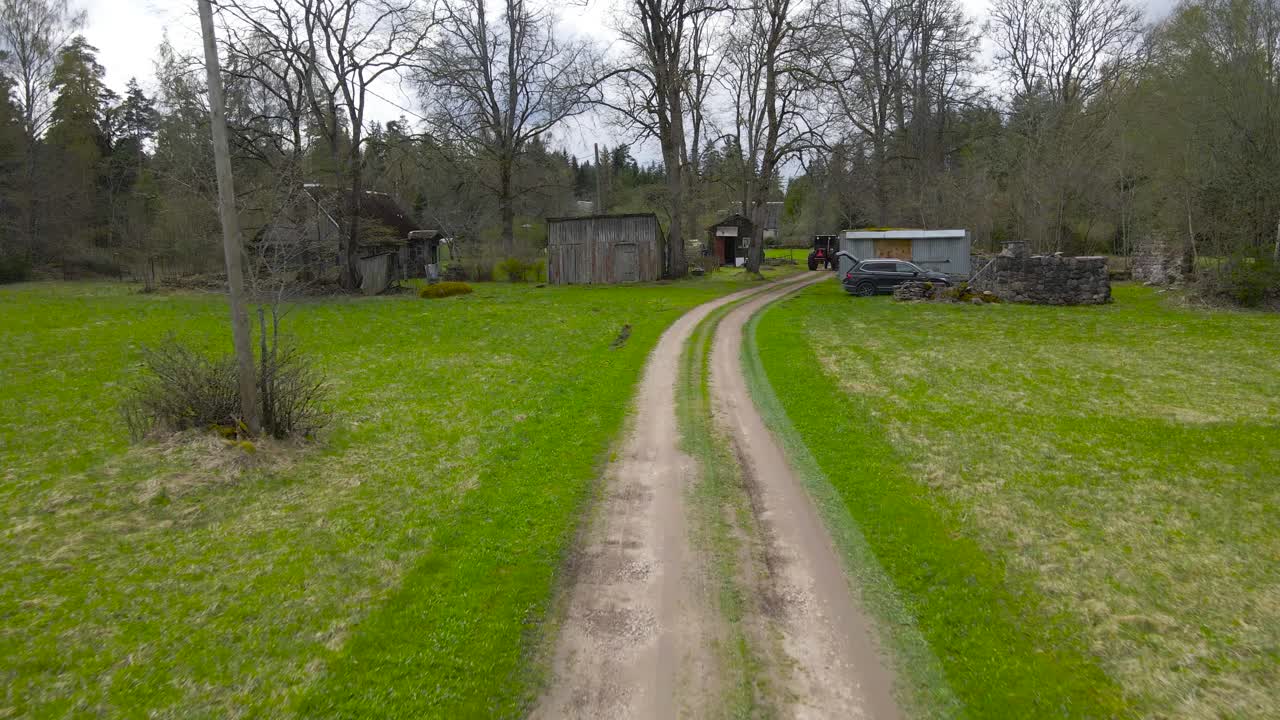 Aerial drone flying and gliding low over a dusty rural countryside muddy brown road that is leading into a garden where people are working with a tractor to spring clean leaves and fallen branches.