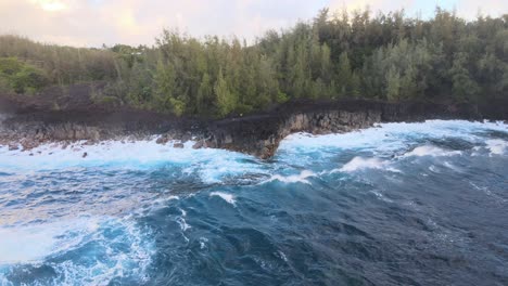 A-drone-gracefully-circles-a-small-cliff-on-the-Big-Island,-Hawaii,-capturing-the-mesmerizing-spectacle-of-deep-blue-waves-pounding-against-the-rugged-shoreline