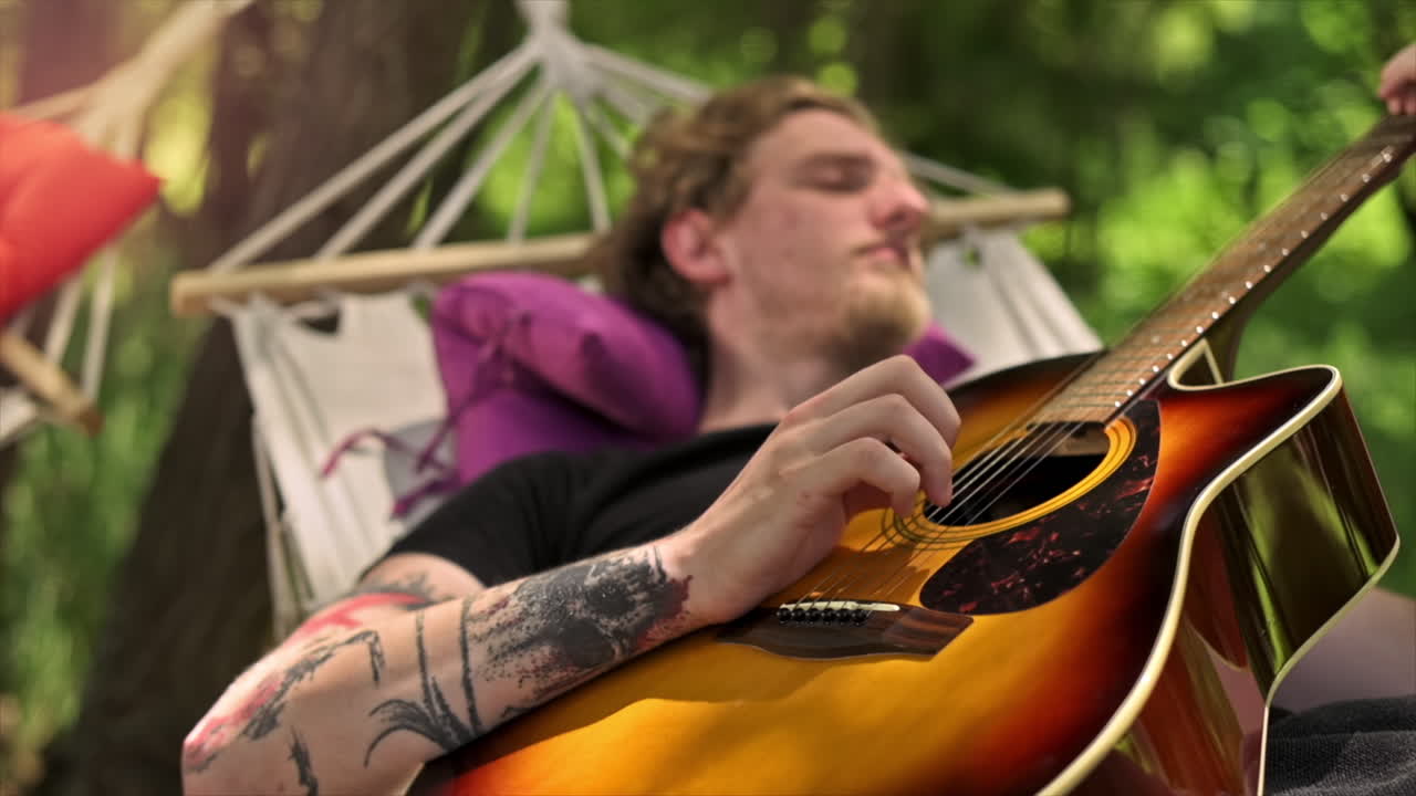 Un giovane sdraiato su un’amaca suona la chitarra. Vegetazione circostante. Glamping. Slow motion