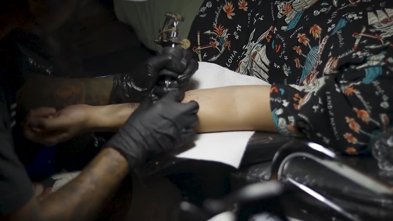 Primo piano di un uomo irriconoscibile a cui viene tatuato un disegno sul braccio, con l'artista che usa una macchinetta per tatuaggi per imprimere l'inchiostro nero nella sua pelle