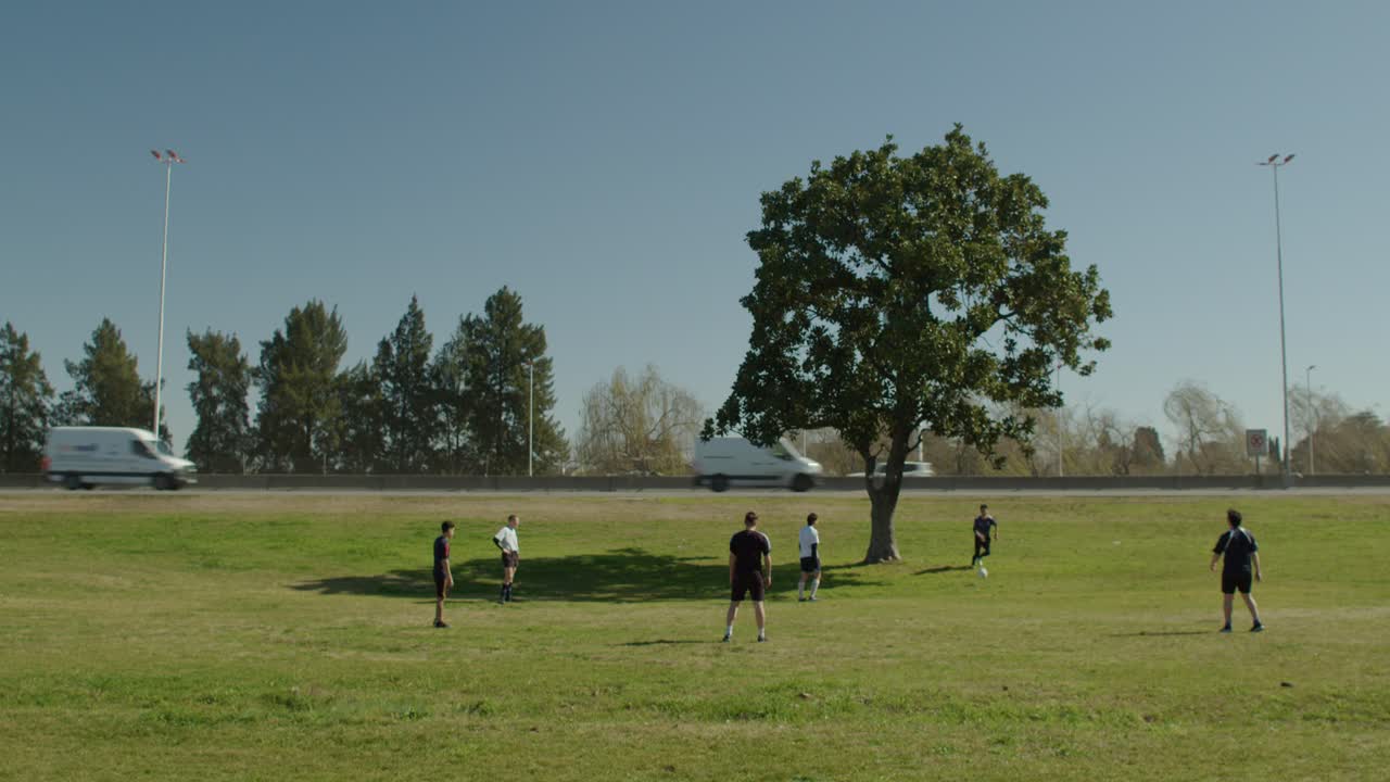 Friends play football soccer on a sunny day in a park near a highway with trucks and trees in the background