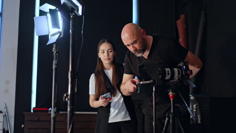 caucasian man stands near his camera on tripod and checking the phone attached to it for prompter. lady with long hair stands behind with phone in hands.