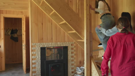 three young women inside wooden cabin kitchen look toward staircase as one climbs up, others observe with interest, natural daylight brightens rustic space with timber walls, visible logs