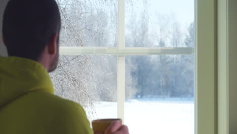 hombre bebe té caliente o café mirando el paisaje de invierno a través de la ventana, cambiando el concepto de enfoque, de cerca