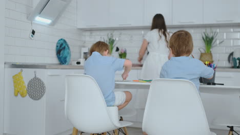 mom in the kitchen washing dishes and two sons sitting at a desk drawing on paper with colored pencils.