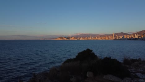 Ascending-over-a-mound-to-reveal-the-tourist-city-of-Benidorm,-Spain---rising-aerial-view