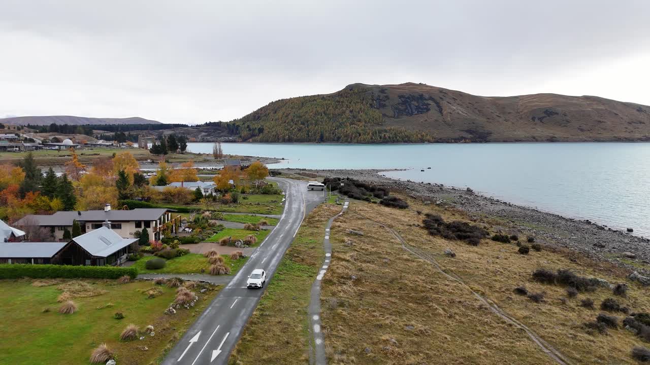Aerial view of a winding road beside Lake Tekapo, showcasing autumn foliage and serene waters under overcast skies