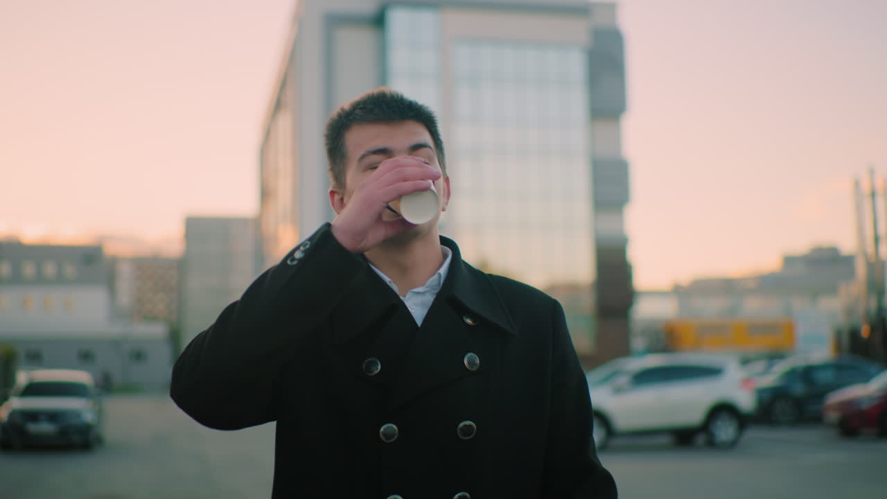 저녁에 바에서 옷을 입은 사업가, 자신감 넘치는 표정으로 커피를 마시고, 배경에는 건물, 주차된 자동차 및 배경의 건물에 빛의 따뜻한 반임으로 구조물이 있습니다.