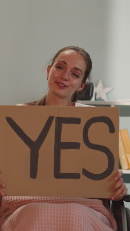 positive woman with disability smiles holding poster with word yes to free access. female person with spine injury sits in wheelchair in room looking in camera