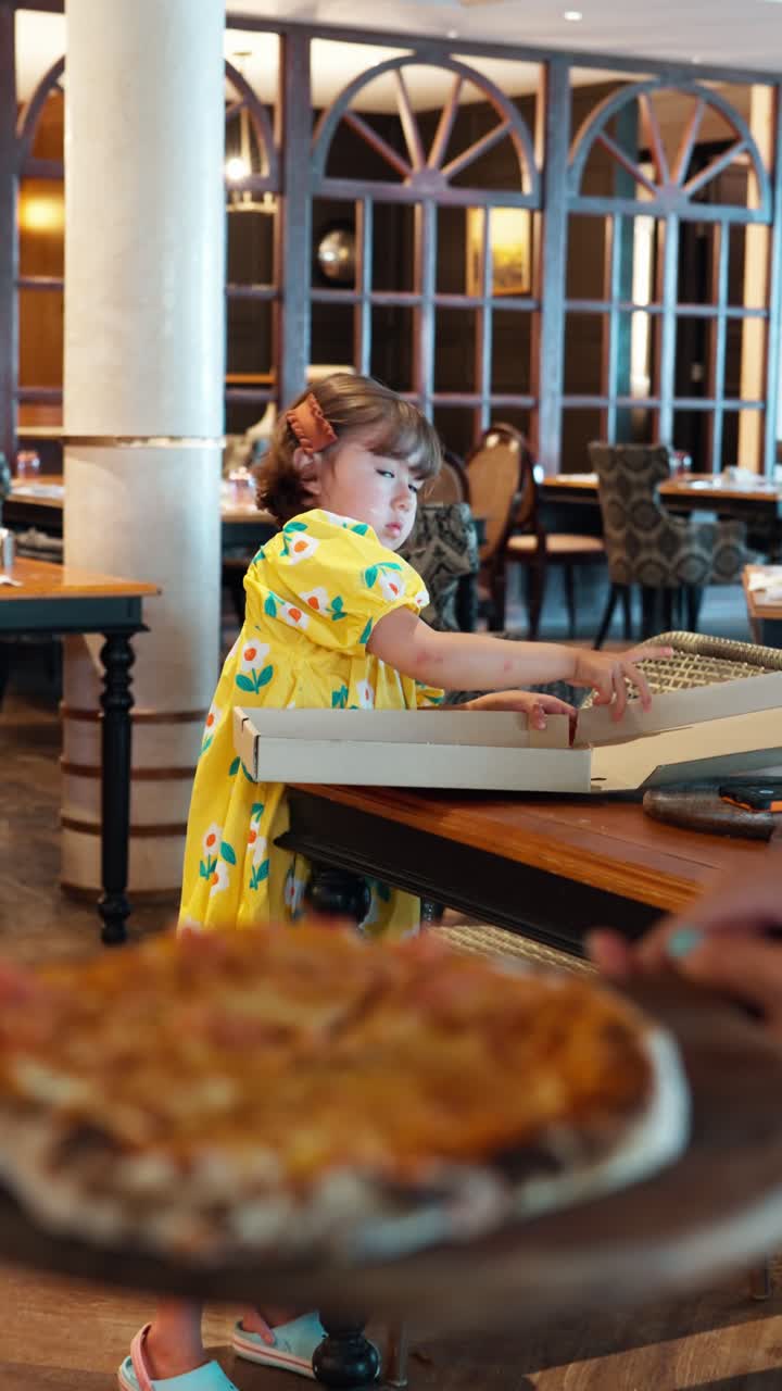 Una bambina carina con un vestito a fiori giallo è in piedi a un tavolo di un ristorante e apre curiosa un grande cartone della pizza per confezionare una pizza appena fatta