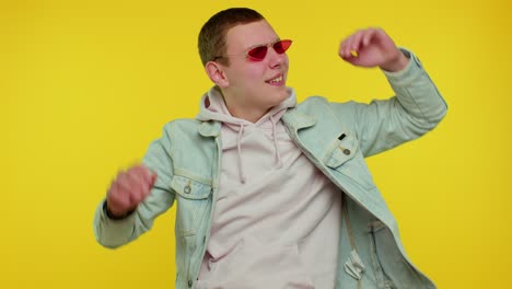 Sincere-cool-cheerful-stylish-man-in-denim-jacket-wearing-sunglasses,-charming-smile-on-yellow-wall