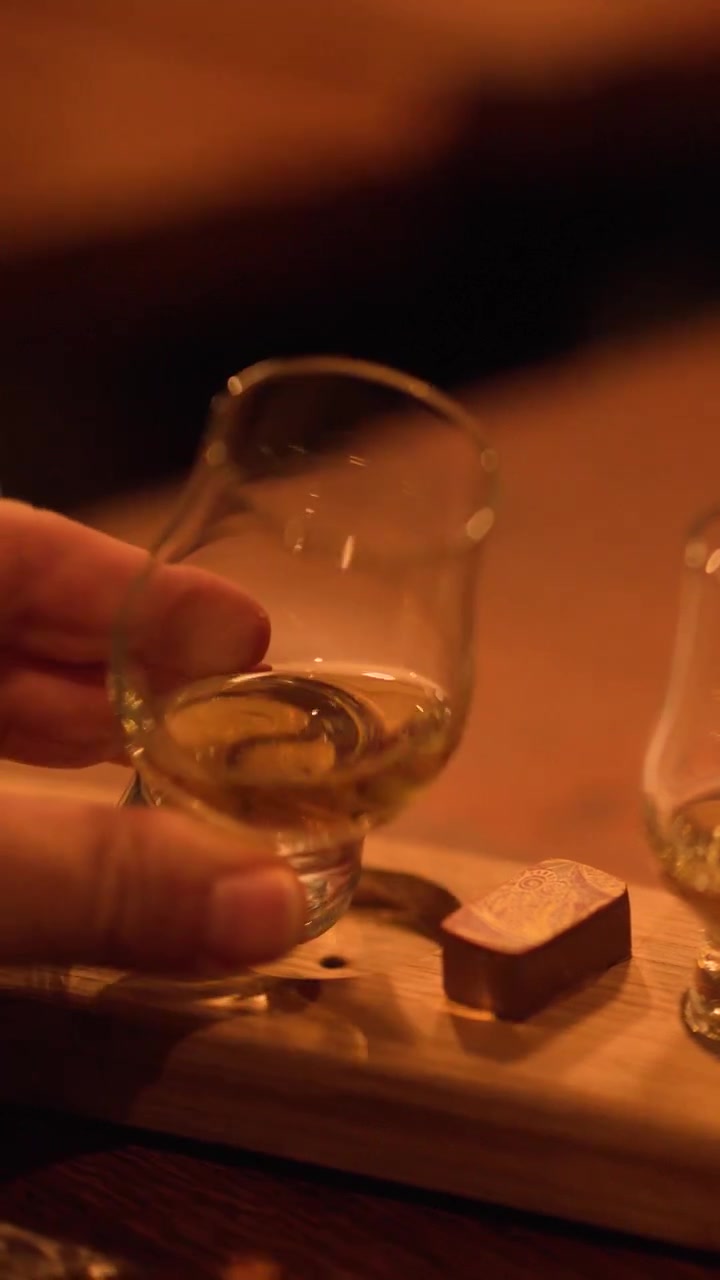 Mano solleva bicchiere di whisky da un tagliere da degustazione sotto un'illuminazione calda, profondità di campo ridotta, primo piano.