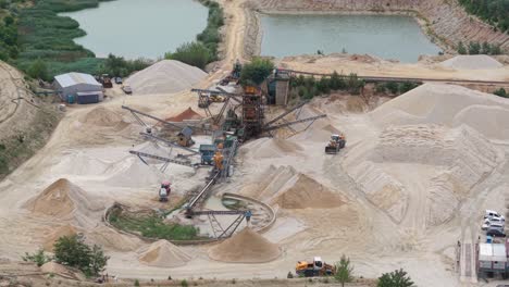 a sand processing facility featuring multiple conveyor belts, large piles of sand and gravel, and heavy machinery. the site is located near two water reservoirs and surrounded by natural vegetation