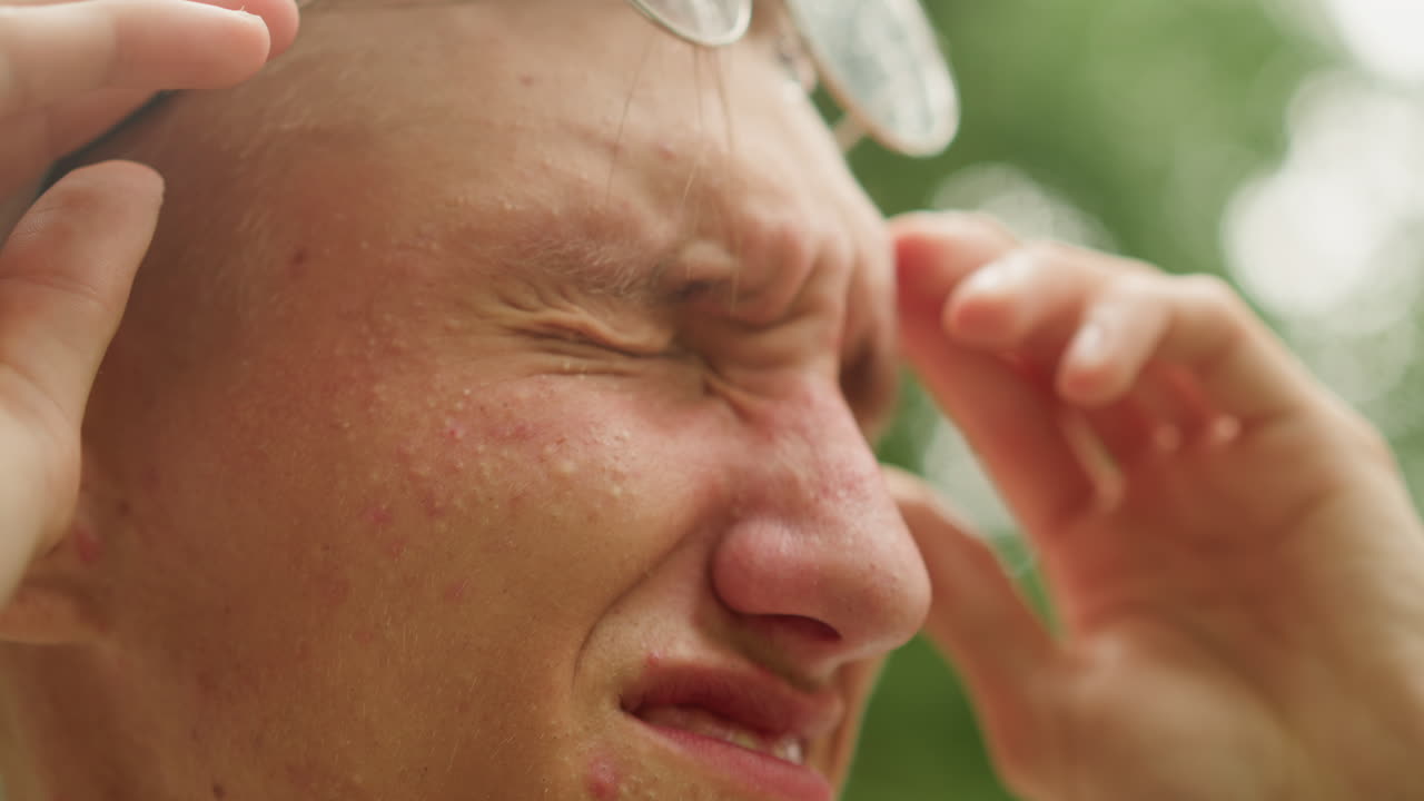 눈물을 닦고 젖은 안경을 고쳐 쓰는 젊은 백인 남성, 빗방울이 맺힌 동그란 렌즈가 있는 야외 클로즈업, 찡그린 얼굴이 안도감으로 풀리고, 손으로 안경테를 안정시키고, 초록색 배경, 봉사자