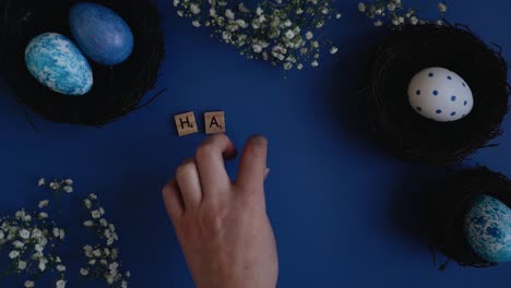 top view of hands writing happy easter with the help of scrabble letters on a blue background surrounded by beautifully decorated easter eggs