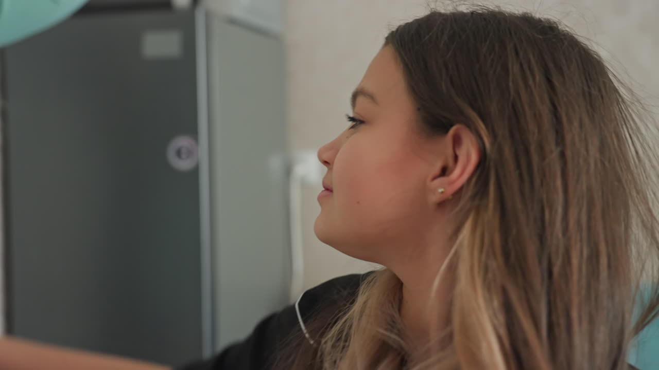 college girl collects spoon from table and pokes friend on head in playful kitchen scene near snack bags and potted plant sisters share laughter cozy indoor moment of quirky bonding over silly game