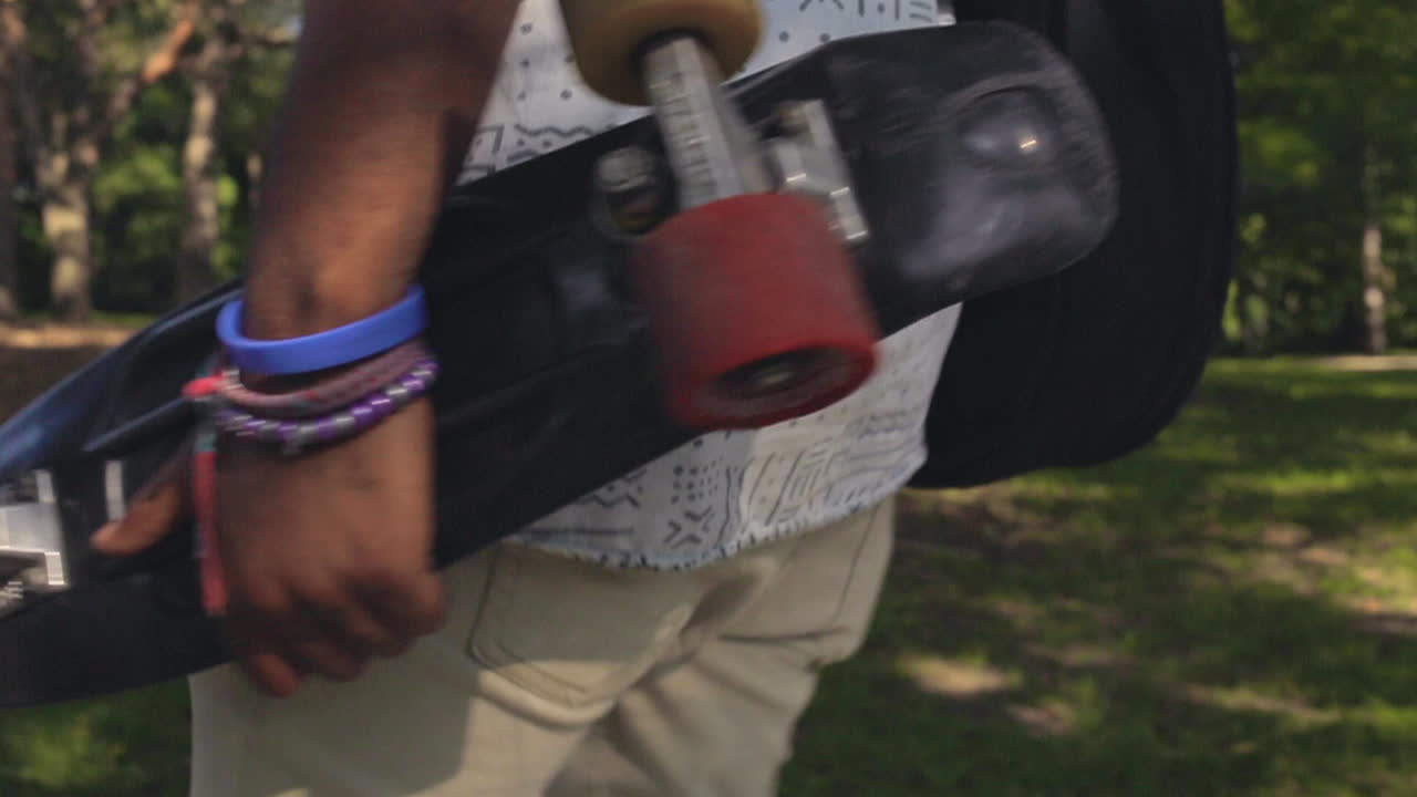 A skateboard cruiser is held in the arm of a black man as he walks through the summertime woods