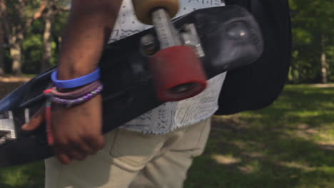 a skateboard cruiser is held in the arm of a black man as he walks through the summertime woods