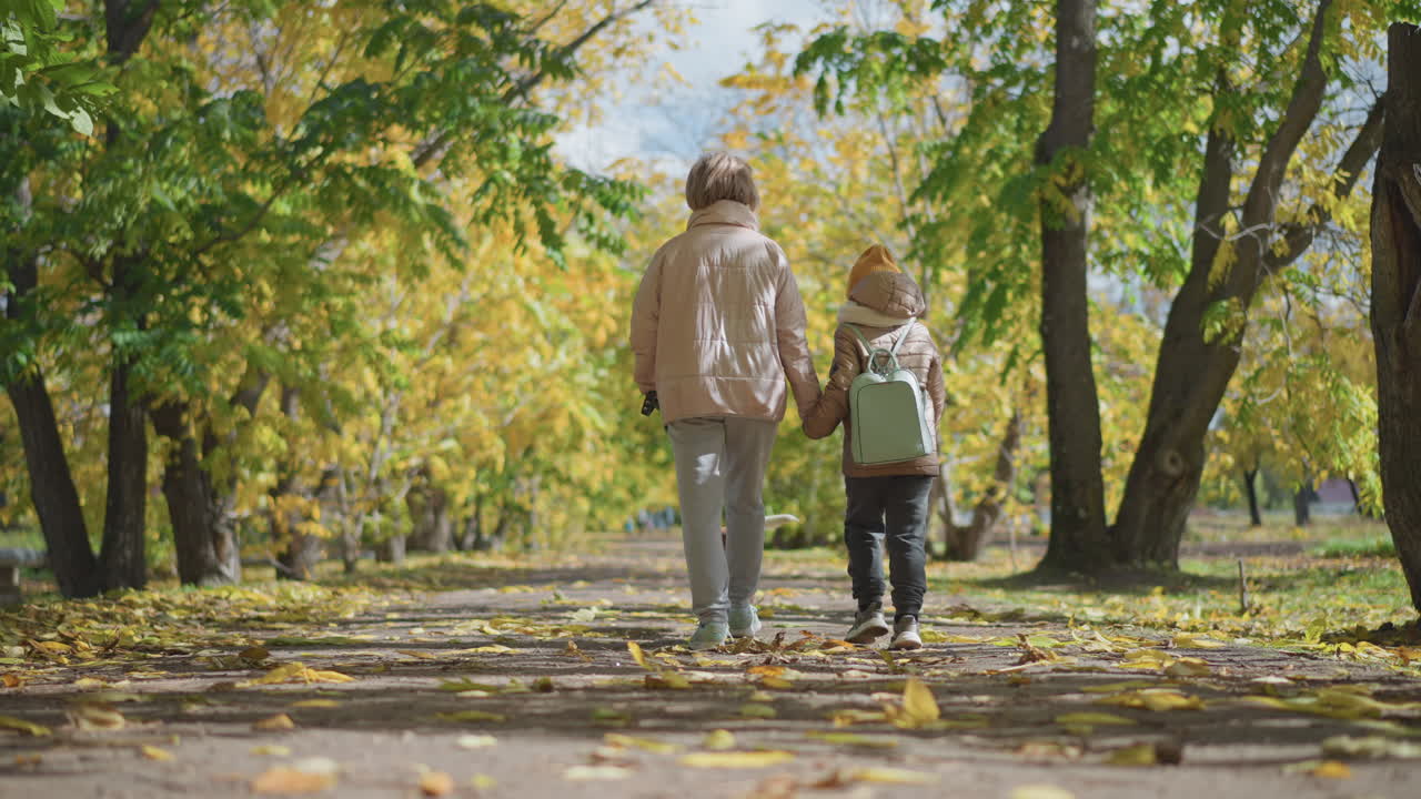 Rear view of child walking with mum as dog trots on leash along leaf strewn autumn forest path under golden canopy, capturing peaceful family stroll, warm sunlit