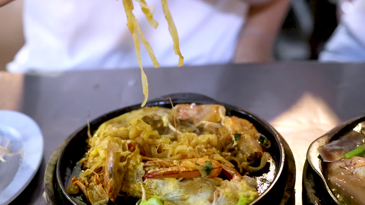 Hand lifts steaming pad thai noodles with shrimp using chopsticks from hot plate in daylight