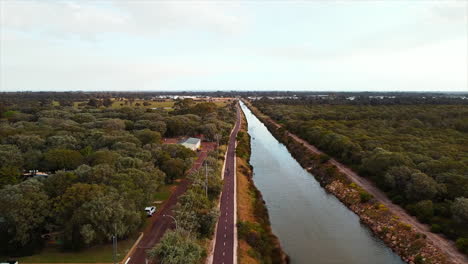 Drohnenansicht-Eines-Flusses,-Der-Im-Flussbett-Fließt,-Und-Eines-Radwegs-Am-Flussufer-Durch-Die-Feuchtgebiete
