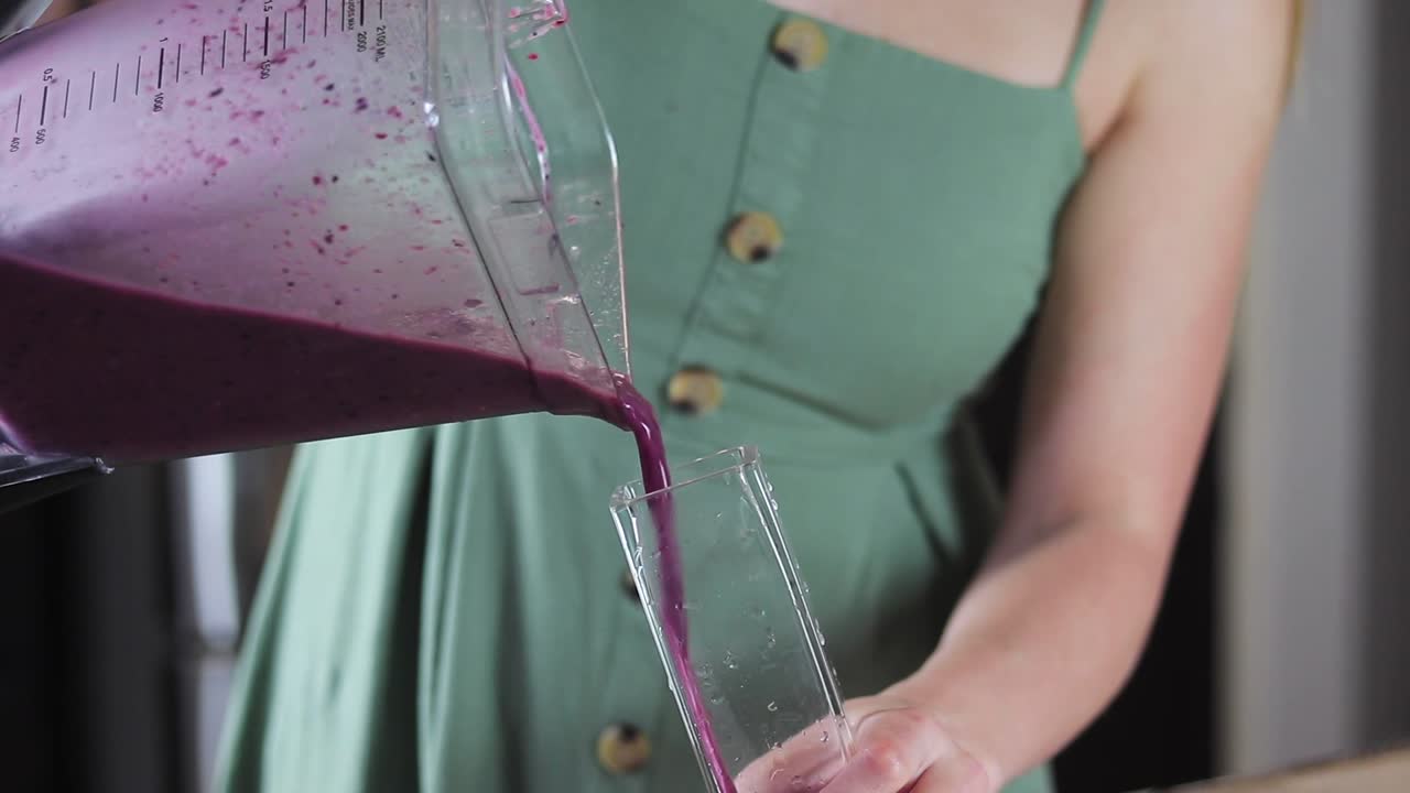 Primo piano al rallentatore di una giovane donna che versa un frullato viola ai mirtilli in un bicchiere di vetro nella sua cucina