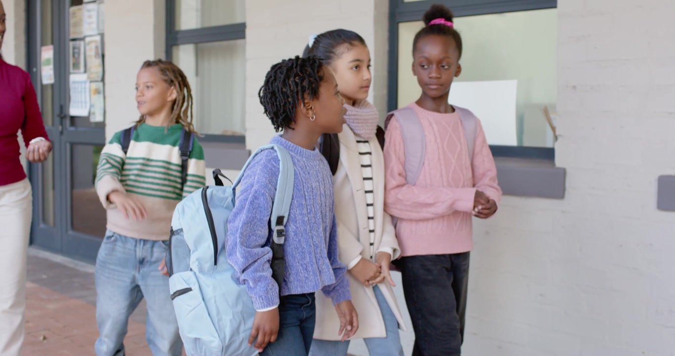 Diverse female pupils walking school path with teacher gesturing, girls stopping to view bulletin